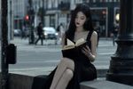 k-pop music video screencap. A girl sits on a low concrete wall near the intersection, alone. She's not in a school uniform. Instead, she wears an elegant black dress that seems out of place on a random Tuesday afternoon—more suited to a banquet than a street corner. Her hair is long, jet-black, falling perfectly straight past her shoulders. Her posture is immaculate. She sits with her legs crossed, reading a leather-bound book with gilt-edged pages, utterly unbothered by the noise around her.

The girl turns a page. Her face is striking—porcelain skin, sharp cheekbones, dark eyes that seem to absorb light rather than reflect it. She carries herself with an effortless, almost terrifying composure.  something about her presence feels ancient. Heavy.
