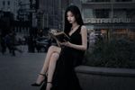 k-pop music video screencap. A girl sits on a low concrete wall near the intersection, alone. She's not in a school uniform. Instead, she wears an elegant black dress that seems out of place on a random Tuesday afternoon—more suited to a banquet than a street corner. Her hair is long, jet-black, falling perfectly straight past her shoulders. Her posture is immaculate. She sits with her legs crossed, reading a leather-bound book with gilt-edged pages, utterly unbothered by the noise around her.

The girl turns a page. Her face is striking—porcelain skin, sharp cheekbones, dark eyes that seem to absorb light rather than reflect it. She carries herself with an effortless, almost terrifying composure.  something about her presence feels ancient. Heavy.
